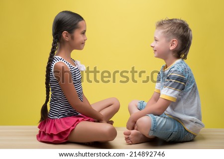 Half-length portrait of pretty dark-haired smiling little girl wearing nice striped dress sitting opposite little fair- haired smiling boy. Isolated on yellow background