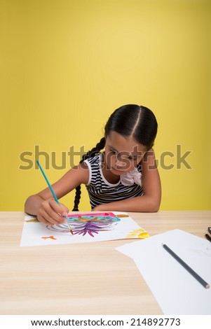 Half-length portrait of pretty dark-haired little girl wearing nice striped dress sitting at the table drawing some nice picture very diligently. Isolated on yellow background