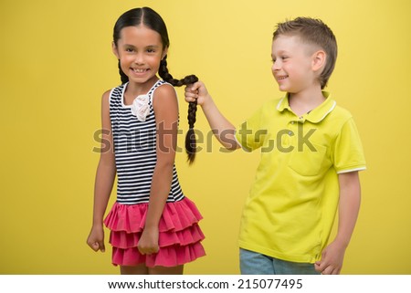 Half-length portrait of lovely little girl wearing nice striped dress and fair-haired smiling boy wearing yellow shirt pulling plait of girl. Isolated on yellow background