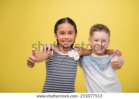 Half-length portrait of lovely little smiling girl wearing nice striped dress and fair-haired smiling boy wearing cool shirt hugging each other showing that everything is great. Isolated on yellow