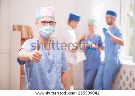 Selective focus on the surgeon wearing blue medical dress showing that everything is OK. His colleagues standing near the window on background