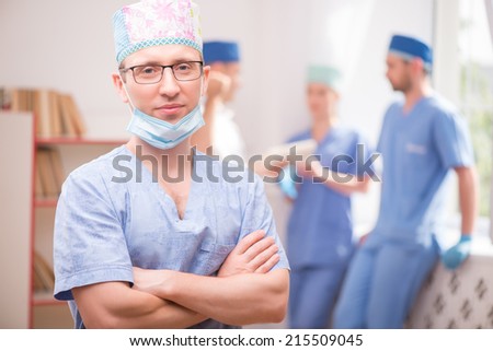 Selective focus on the surgeon wearing blue medical dress crossing his hands standing and looking at us. His colleagues standing near the window on background