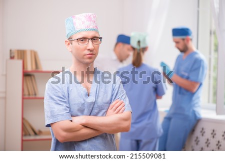 Selective focus on the surgeon wearing blue medical dress crossing his hands standing and looking at us. His colleagues standing near the window on background