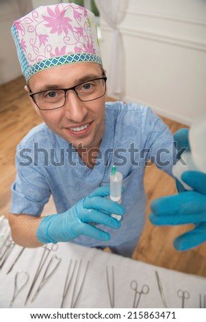 Half-length portrait of the smiling doctor wearing a cap glasses blue medical uniform and disposable gloves standing in the operating room holding a syringe looking at us. Top view