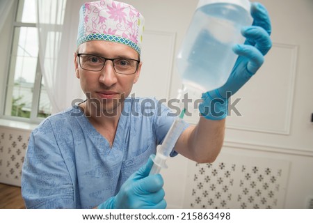 Half-length portrait of the smiling doctor wearing a cap glasses blue medical uniform and disposable gloves standing in the operating room filling up the syringe with the fluid