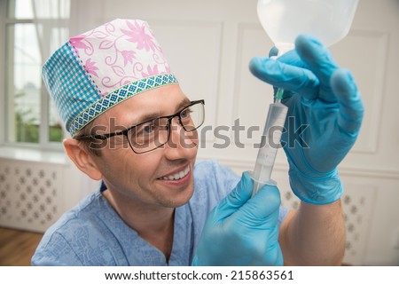 Half-length portrait of the smiling doctor wearing a cap glasses blue medical uniform and disposable gloves standing aside filling up the syringe with some fluid