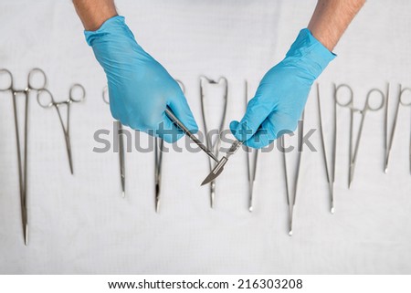 Selective focus on the hands of the doctor wearing blue disposable gloves holding the scalpel over the other medical instruments lying on the white material on background