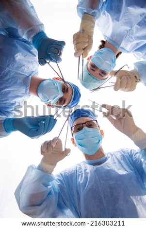 Half-length portrait of three doctors wearing blue medical masks and uniform holding a pair of scissors and tweezers in both hands operating the patient. Low angle