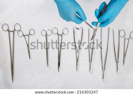 Selective focus on the hands of the doctor wearing blue disposable gloves holding the scalpel over the other medical instruments lying on the white material on background