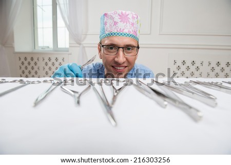 Half-length portrait of the smiling doctor wearing glasses  holding a scalpel looking out of the table with different medical instruments on it