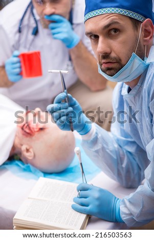 Selective focus on the surgeon sitting in the operation room near the victim holding his cigarette with the help of tweezers looking at us. His colleague drinking coffee on background