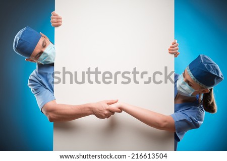 Half-length portrait of two doctors wearing blue medical uniform and masks looking out of the huge white poster for copy place and holding hands. Isolated on blue background