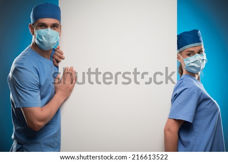 Half-length portrait of two doctors wearing blue medical uniform and masks leaning on the huge white poster for copy place. Isolated on blue background
