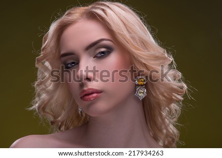 Half-length portrait of beautiful sexy blonde with evening make up wearing great diamond earrings fingering and bracelet putting her hands on her shoulders looking at us seductively isolated on dark