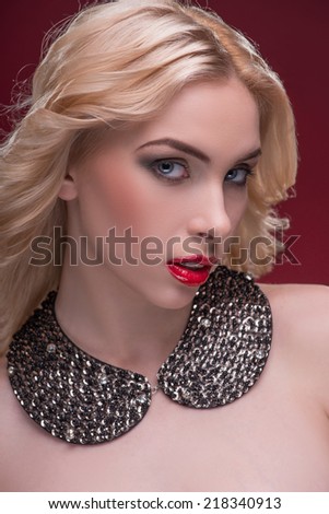 Half-length portrait of beautiful sexy blonde with red tempting lips and perfect skin wearing huge black necklace looking out for someone isolated on dark background