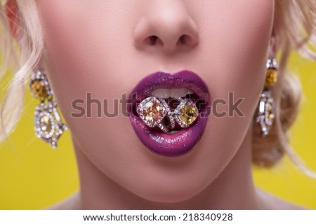 Half-length portrait of beautiful sexy blonde with violet tempting lips and perfect skin keeping two gold fingerings in her mouth isolated on yellow background. Close up