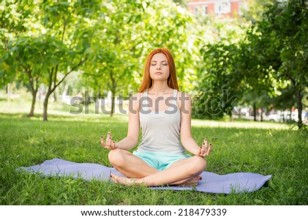 Pretty young red-haired woman wearing white T-shirt and mental shorts sitting on the blue mat doing yoga in the park (lotus pose)