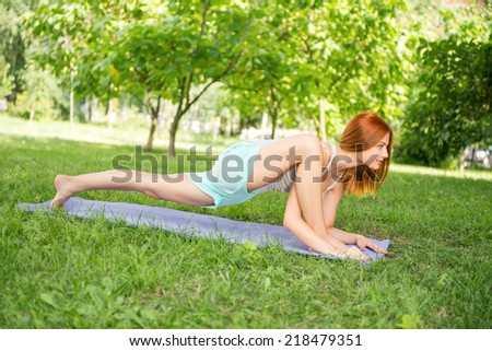 Pretty young red-haired woman wearing white T-shirt and mental shorts doing yoga stretching her legs on the blue mat in the park