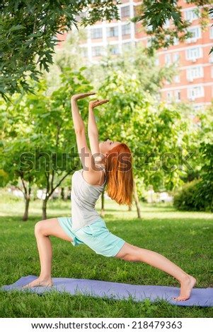 Pretty young red-haired woman wearing white T-shirt and mental shorts doing yoga stretching in different pose  on the blue mat in the park