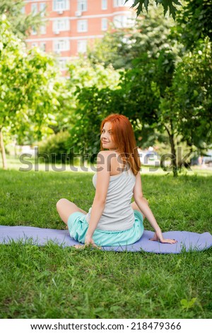Pretty young red-haired woman wearing white T-shirt and mental shorts doing yoga sitting back on the blue mat in the park looking out at us