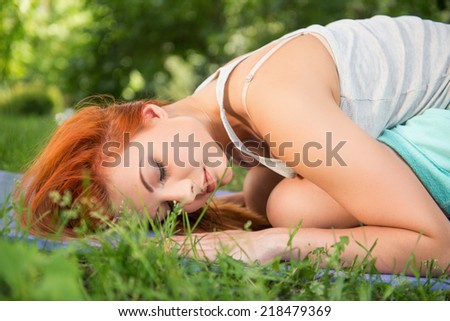 Pretty young red-haired woman wearing white T-shirt and mental shorts doing yoga lying like a ball on the blue mat in the park