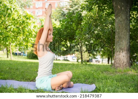 Pretty young red-haired woman wearing white T-shirt and mental shorts doing yoga sitting aside on the blue mat in the park