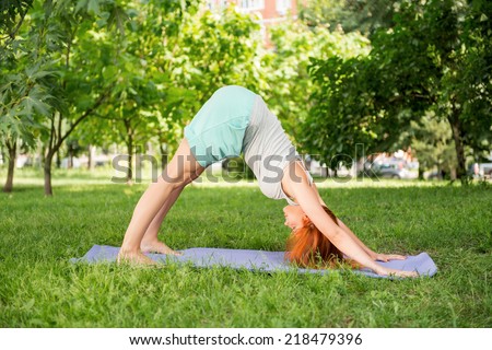 Pretty young red-haired woman wearing white T-shirt and mental shorts doing yoga standing in the pose fanny up on the blue mat in the park