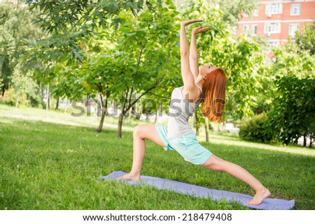 Pretty young red-haired woman wearing white T-shirt and mental shorts doing yoga stretching in different pose  on the blue mat in the park