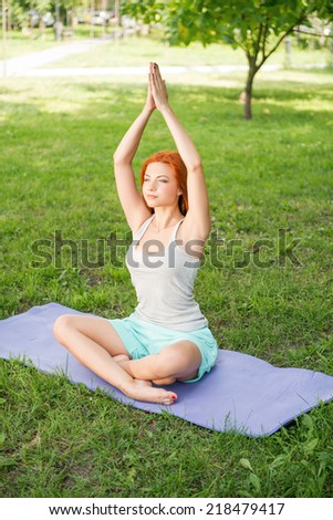 Pretty young red-haired woman wearing white T-shirt and mental shorts doing yoga lotus pose sitting on the blue mat in the park