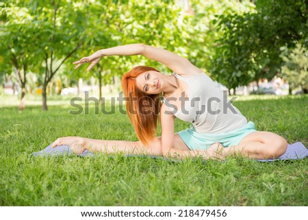 Pretty young red-haired woman wearing white T-shirt and mental shorts doing yoga stretching on the blue mat in the park