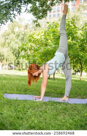 Pretty young red-haired woman wearing white T-shirt and grey pants doing yoga standing on the one hand on blue mat in the park