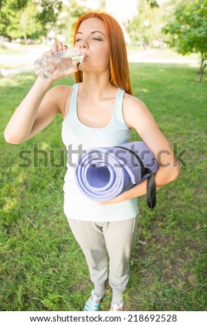Pretty young red-haired woman wearing white T-shirt and grey pants standing in the park drinking water thinking about the beauty of life. Top view