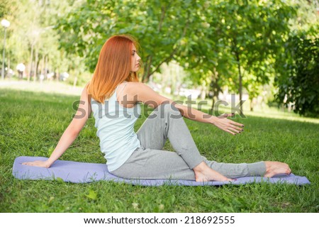 Pretty young red-haired woman wearing white T-shirt and grey pants doing yoga stretching crossing her legs on blue mat in the park