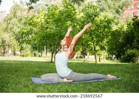 Pretty young red-haired woman wearing white T-shirt and grey pants doing yoga stretching lying on blue mat in the park