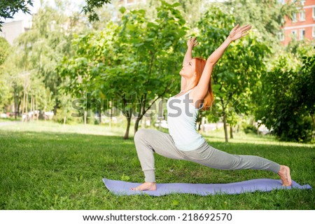 Pretty young red-haired woman wearing white T-shirt and grey pants doing yoga stretching lying on blue mat in the park