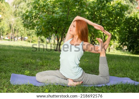 Pretty young red-haired woman wearing white T-shirt and grey pants doing yoga stretching her legs and hands sitting on blue mat in the park
