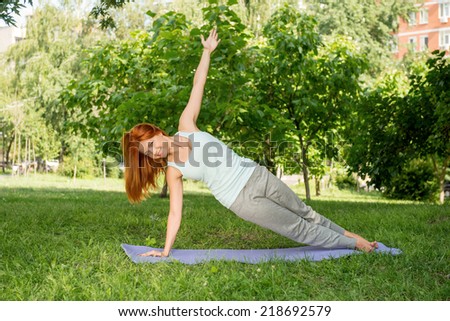 Pretty young red-haired woman wearing white T-shirt and grey pants doing yoga pressing out of the ground on blue mat in the park