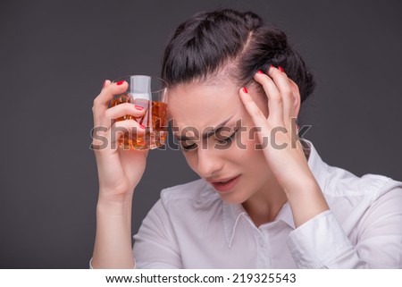 Half-length portrait of dark-haired tired beautiful woman wearing white blouse standing aside holding a glass of whisky touching her head looking at us. Isolated on dark background