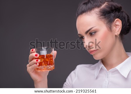 Half-length portrait of dark-haired beautiful woman wearing white blouse standing aside holding a glass of whisky looking at it. Isolated on dark background