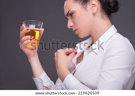 Half-length portrait of dark-haired beautiful woman wearing white blouse sitting aside holding a glass of beer unfastening her blouse. Isolated on dark background