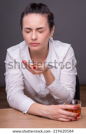 Half-length portrait of dark-haired beautiful desperate woman wearing white blouse sitting at the table closing her eyes holding a glass of whisky wanted to swallow a pile of colorful pills. Isolated