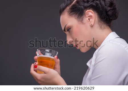 Half-length portrait of dark-haired beautiful woman wearing white blouse sitting aside holding a glass of whisky and looking at it. Isolated on dark background