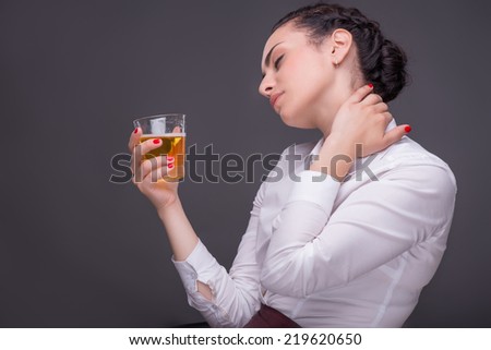 Half-length portrait of dark-haired beautiful woman wearing white blouse sitting aside with closed eyes holding a glass of whisky crumbling her neck. Isolated on dark background