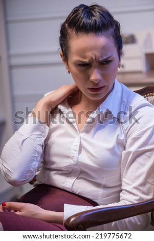 tired woman wearing white blouse sitting in the chair crumbling her neck and thinking how to solve her problems. Her office on background