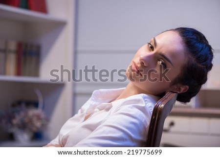 woman wearing white blouse and vinous skirt sitting in the chair looking at us with despair in her eyes. Her office on background
