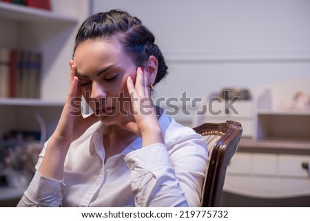 young tired woman wearing white blouse sitting in the chair with closed eyes holding at her head and thinking hard about big problems in her company. Her