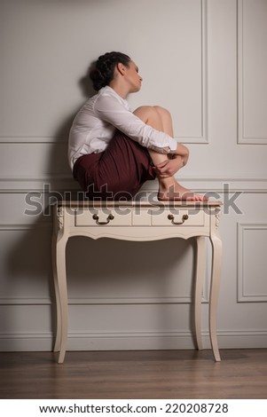 Full-length portrait of beautiful dark-haired fagged young woman wearing white blouse and vinous skirt sitting on the little table leaning to the wall thinking about her husband
