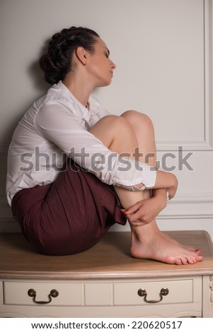 Full-length portrait of tired business woman wearing white blouse and vinous skirt sitting on the table with closed eyes leaning to the wall