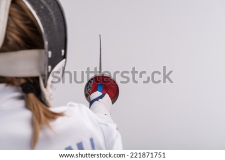 Selective focus on the sword in the hands of woman wearing white fencing costume and black fencing mask standing back to us. Isolated on white background