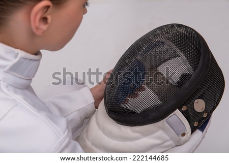 Half-length portrait of pretty young woman wearing white fencing costume sitting aside holding and looking at her black fencing mask in her hands. Isolated on white background